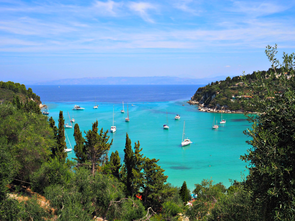 Bright clear turquoise sea shading to darker azure in a bay surrounded by green hillsides on Paxos island Greece. Copyright@2024 mapandfamily.com