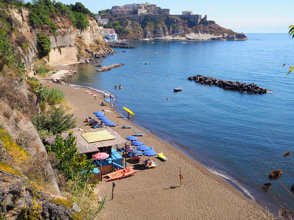 Day trip to Procida. View from stairs to dark sand beach below with blue sun umbrellas. Copyright©2019 mapandfamily.com 