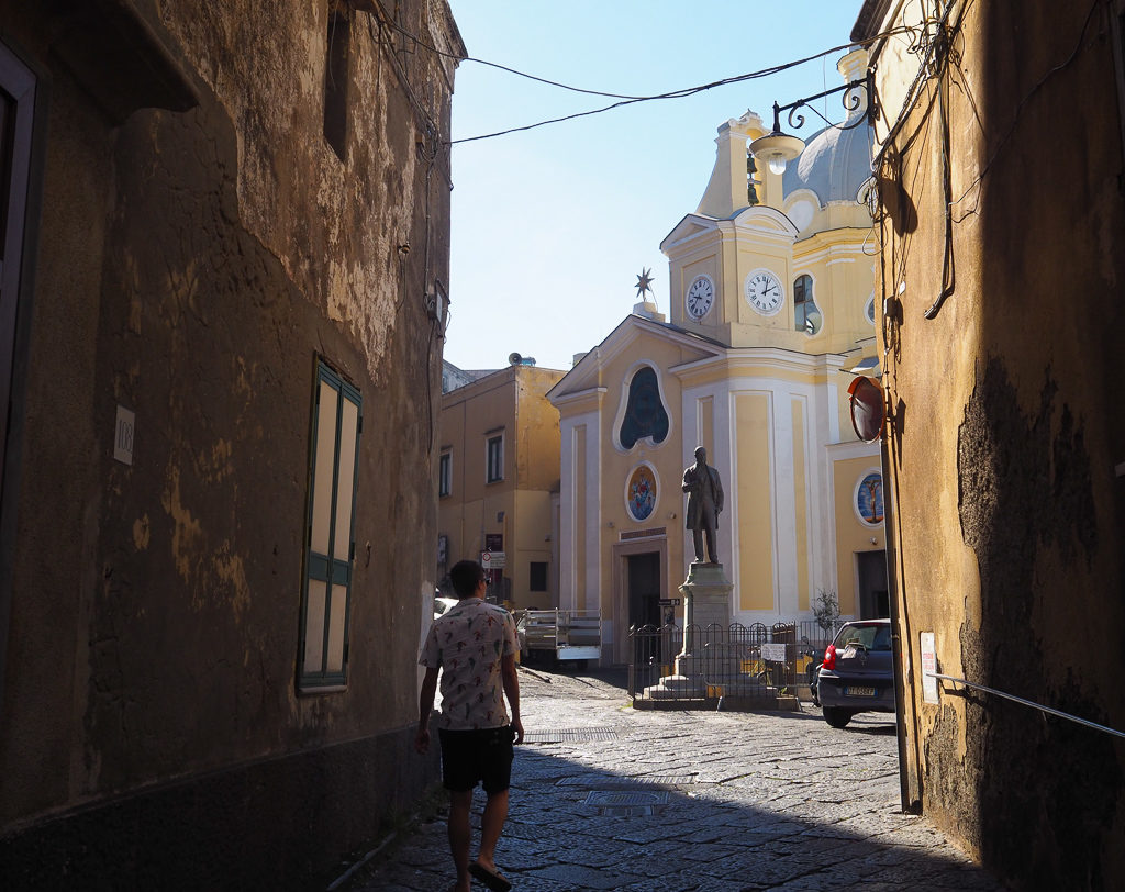 Day trip to Procida island. Boy walking from narrow street into square with church and statue. Copyright©2019 mapandfamily.com 