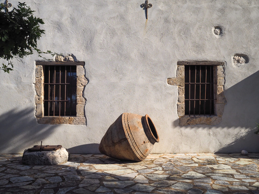 Villa in Kefalonia. Exterior wall of restored olive mill with original metal barred windows and urn on paved terrace. Copyright ©2020 mapandfamily.com