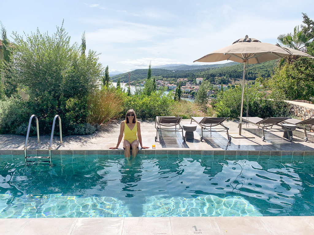 Fiskardo Kefalonia, woman in yellow swimsuit sitting by pool with bay in background. Copyright ©2020 mapandfamily.com 