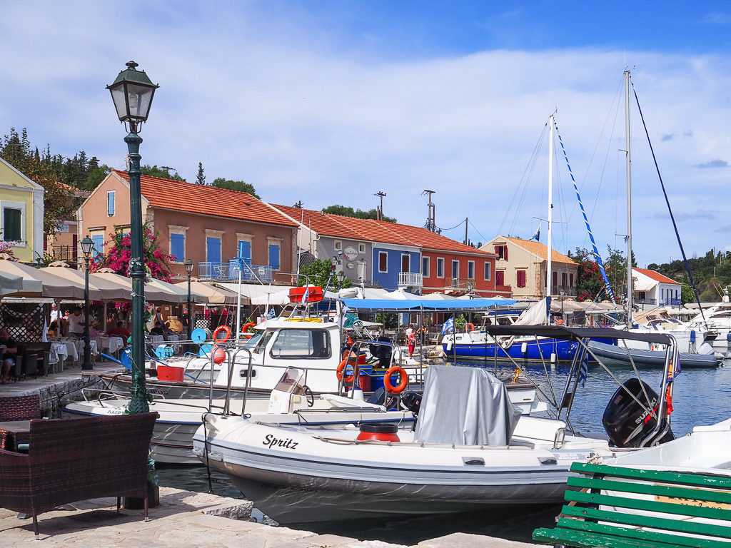 Fiskardo village, view of the waterfront with green lamppost and boats moored up. Coloured houses overlook the water. Copyright ©2020 mapandfamily.com 