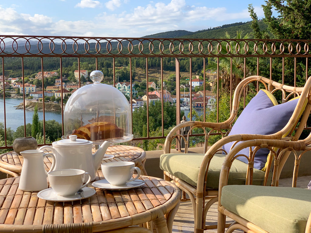 Fiskardo villas. Low table and chairs with cushions on balcony with teapot, cups and a homemade cake. Fiscardo village in background. Copyright ©2020 mapandfamily.com 