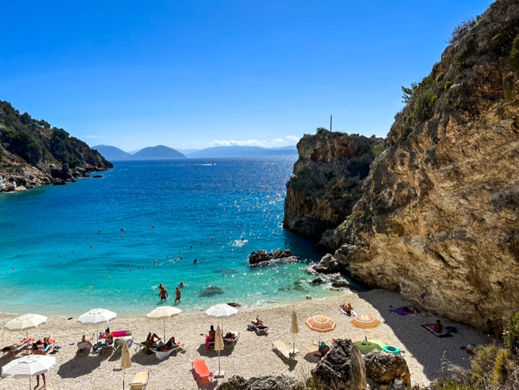 Looking down on to Agiofili beach near Vasiliki Lefkada. People paddling in shallow turquoise water. Others sitting under parasols. Copyright@ 2022 mapandfamily.com 