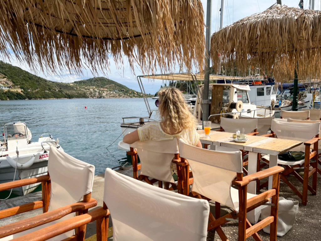 Woman sitting at a quayside table with a glass of orange juice. Sivota, Lefkada. Copyright@ 2022 mapandfamily.com 