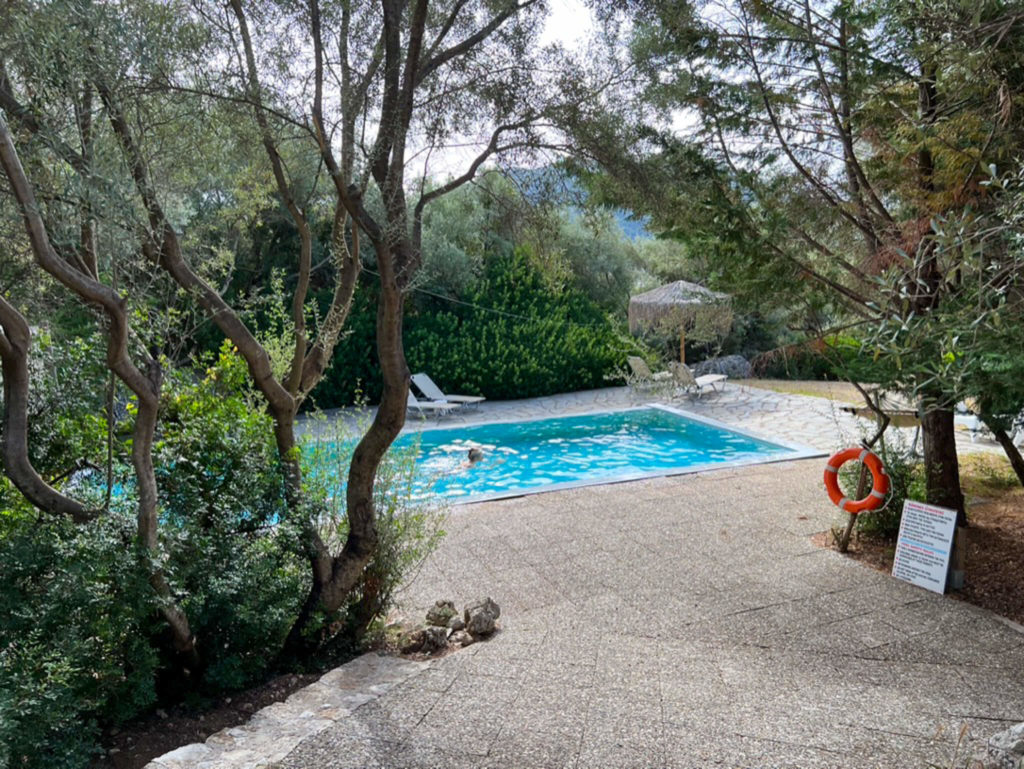 Woman swimming in pool in garden of Thimari villa in Vasiliki. Orange lifebelt on tree, sun loungers and parasol beside pool. Copyright@ 2022 mapandfamily.com 