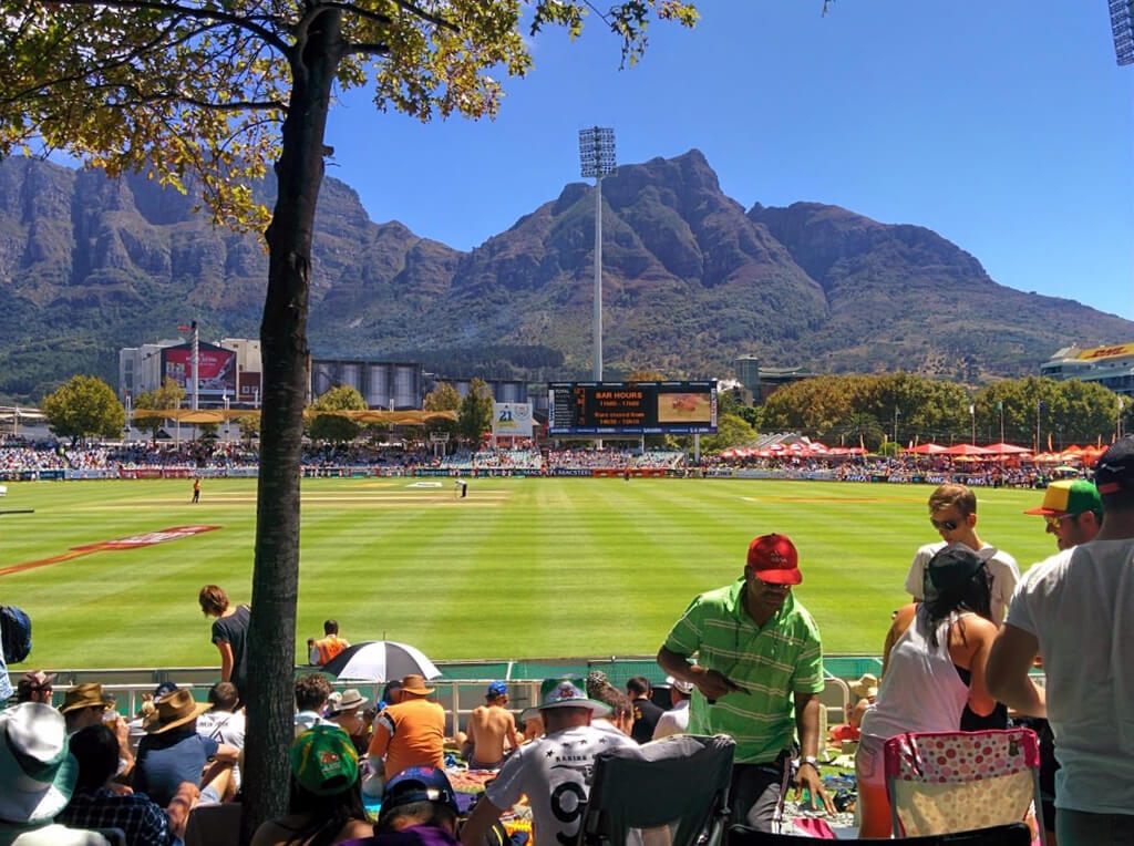 A green cricket pitch with mountains behind it, the audience in the foreground are sitting on blankets. Copyright@2023 reserved to photographer via mapandfamily.com