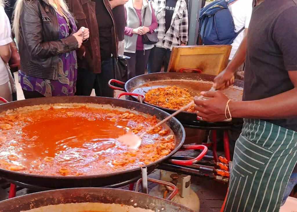 A server stirring a large pan of food whilst customers watch at a food stall in a market. Copyright@2023 reserved to photographer via mapandfamily.com