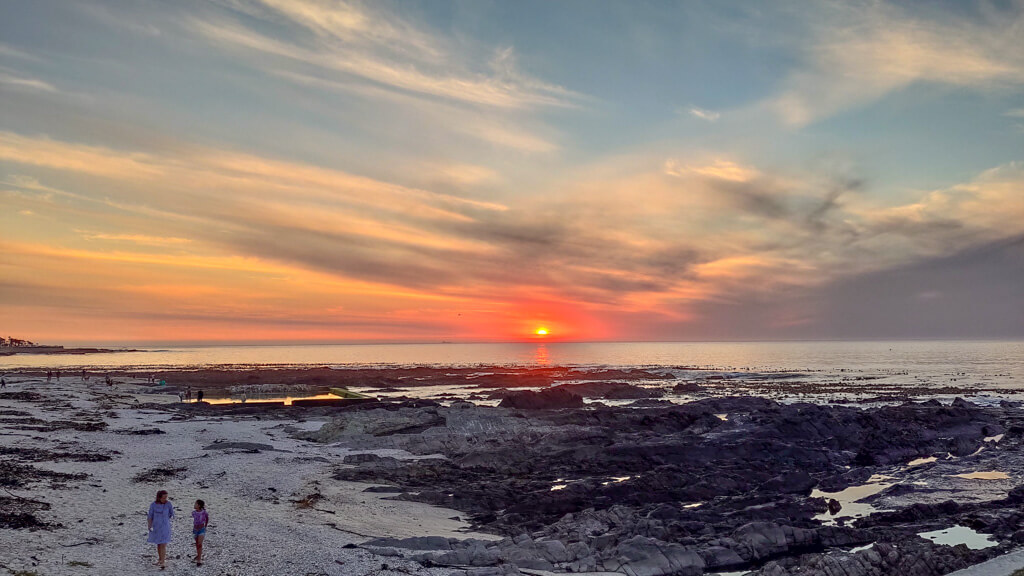 Sun setting into sea on horizon. Rocky beach in foreground with two figures on it. Copyright@2023 reserved to photographer via mapandfamily.com 