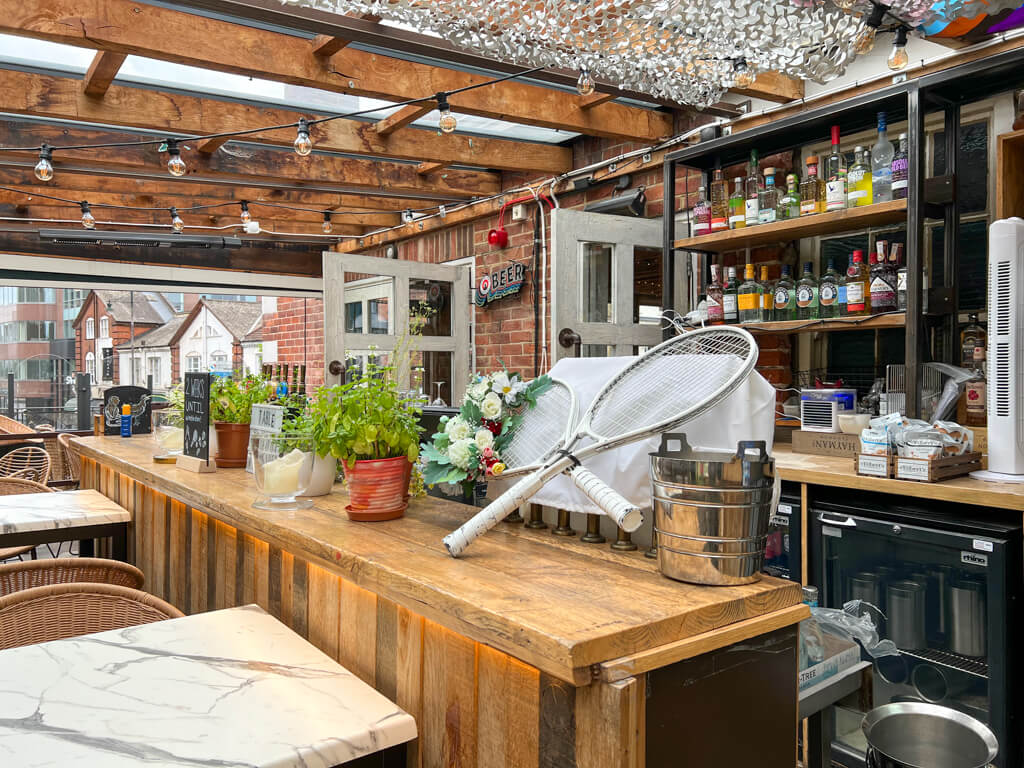 Covered roof terrace with bar, tennis racket decor, strings of lights and tables in foreground. Copyright@2023mapandfamily.com 