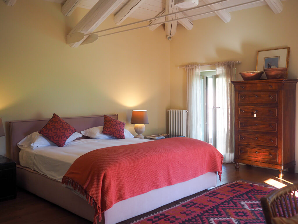 Villa Giara bedroom with dark red throw on bed, toning cushions, dresser and french doors. Copyright@2023mapandfamily.com