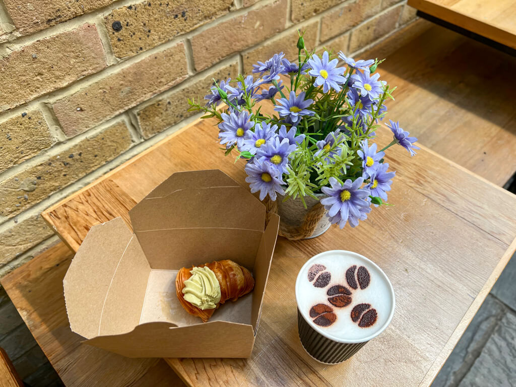 Table on patio of Wimbledon cafe with capuccino in a paper cup with coffee bean design in chocolate on top. Italian pastry in little card box and small vase with decorative blue flowers. Copyright@2023 mapandfamily.com 