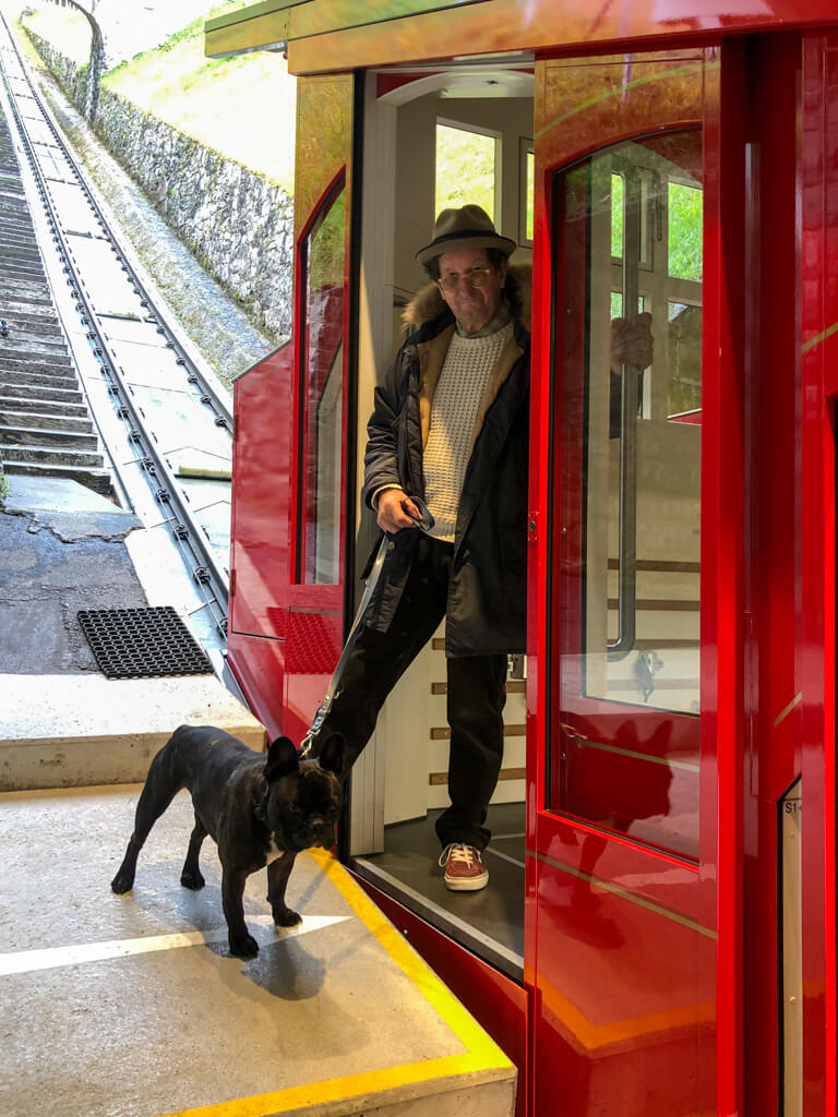 Man wearing a hat with French bulldog step out of red painted furnicular lift. Copyright@2023 reserved to photographer via mapandfamily.com 