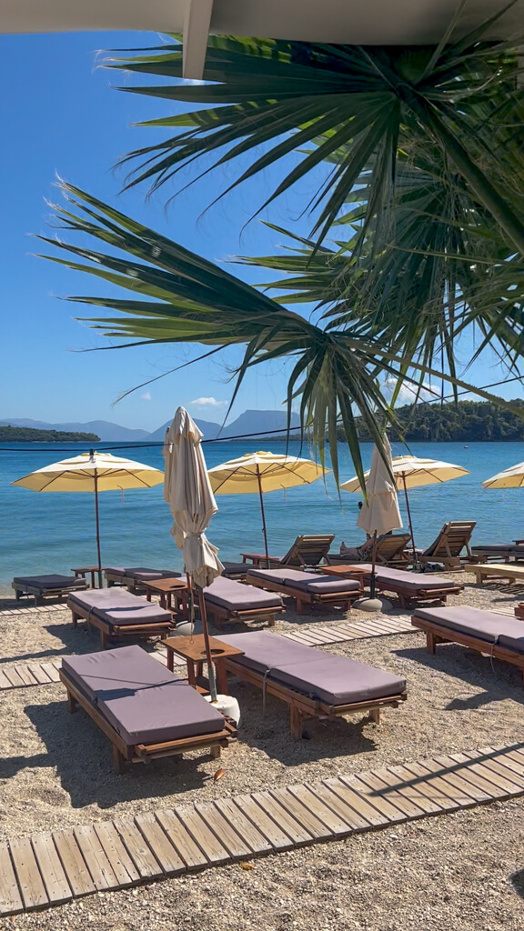 Sun loungers, parasols and palm fronds with a blue bay in background at Sailinn beach club, Lefkada.Copyright@2023mapandfamily.com 