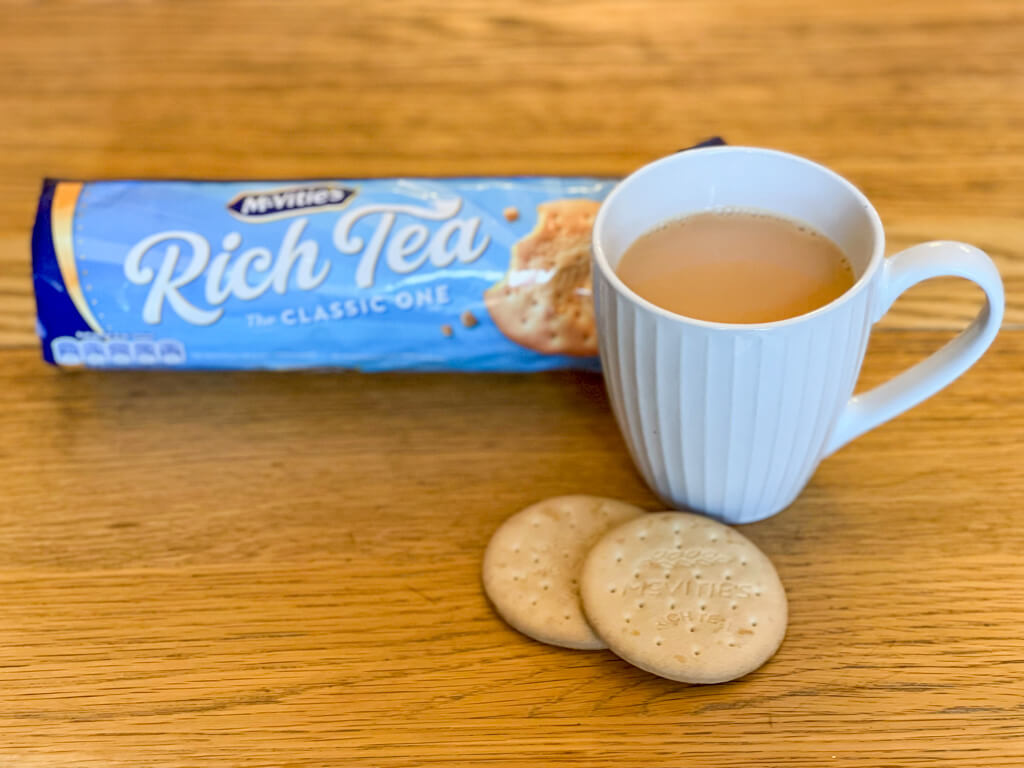 A cup of tea in a white fluted mug with two Rich Tea biscuits and a packet in the background. Copyright 2023@mapandfamily.xyz