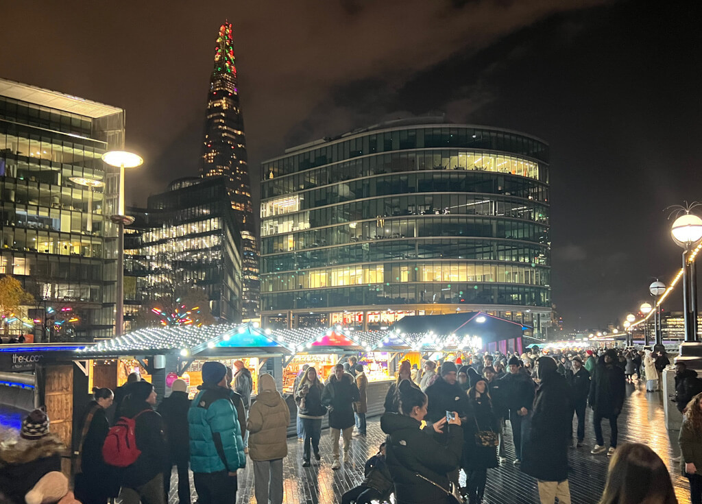 Evening at London Bridge Christmas market as crowds gather around illuminated stalls, with Shard in background. Copyright@2023 mapanfamily.com 