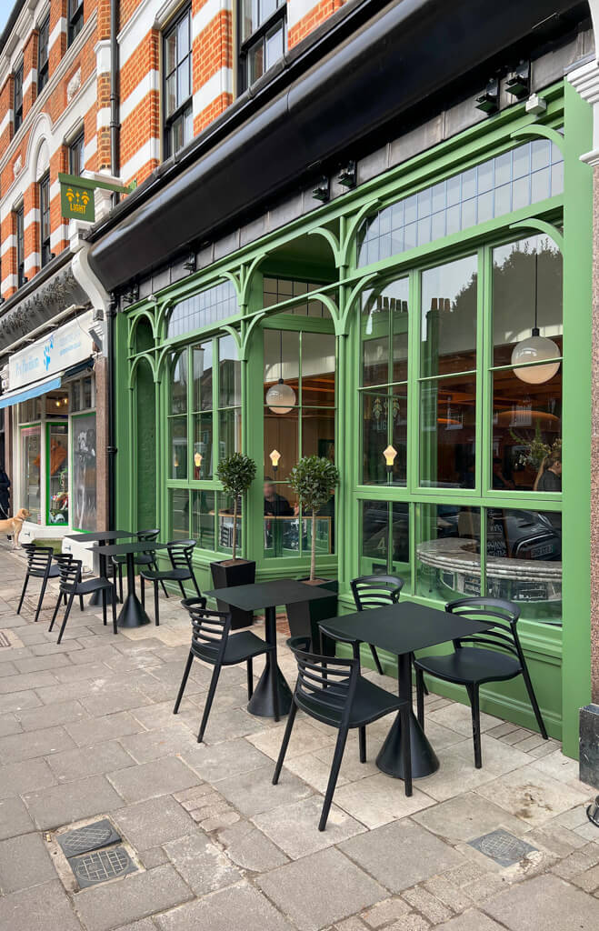 Black tables and chairs on pavement outside the green painted and glazed facade of Light on the Common. Copyright@2024 Nancy Roberts