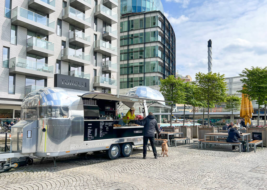 Silver Airstreem trailer serving coffees and snacks. A customer and dog stand beside it and alongside are benches and trees beside the canal. Copyright@2024 mapandfamily.com