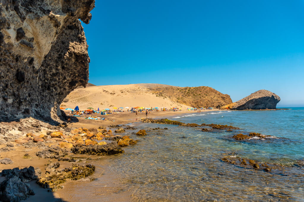 Lava rocks on the natural beach of Monsul with golden sand and people sitting under parasols at the water's edge. 