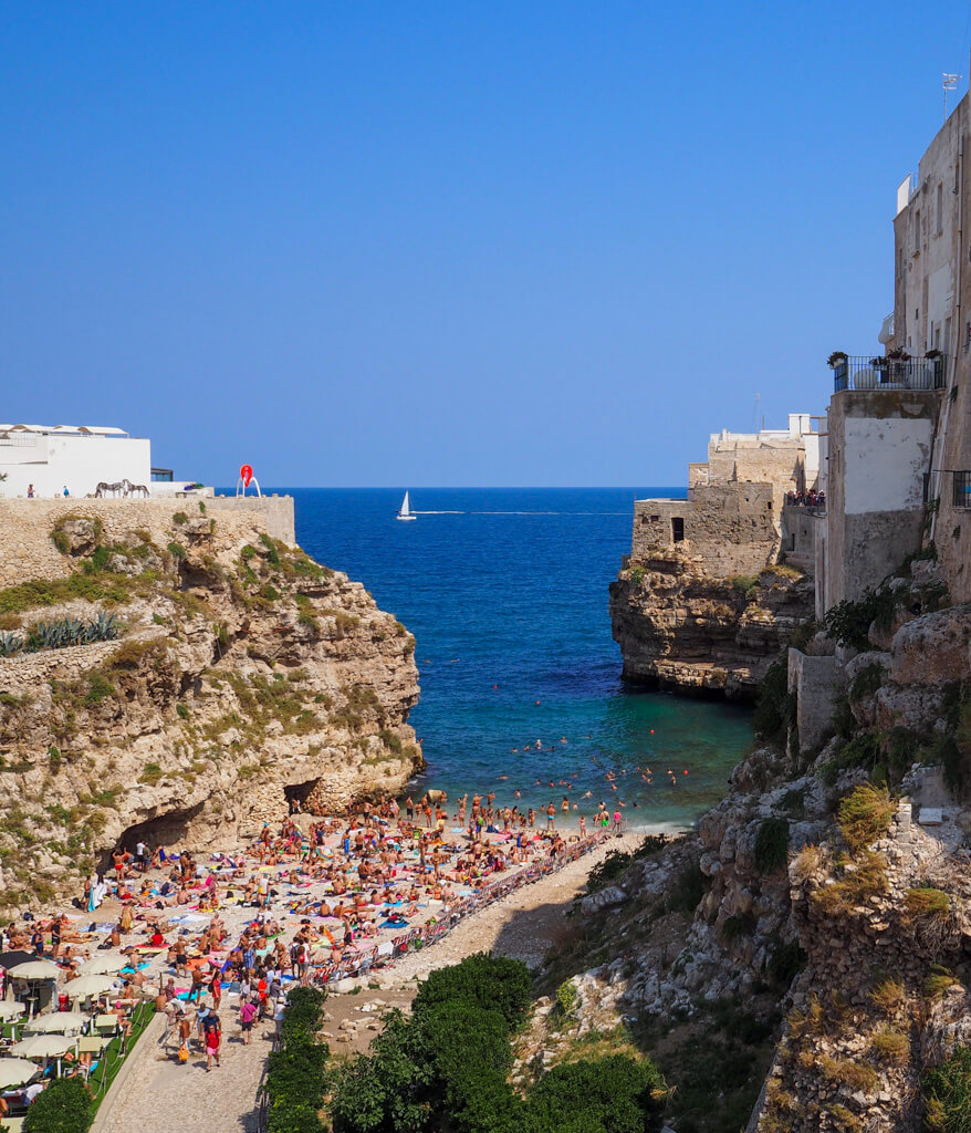 A busy pebbly beach with high cliffs on either side and deep blue sea at Polignano a Mare. Copyright@2024 mapandfamily.com 