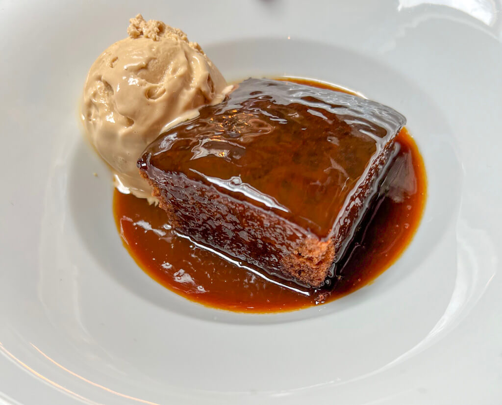 A glossy portion of sticky toffee pudding on a white plate with a scoop of ice cream. Copyright@2024NancyRoberts